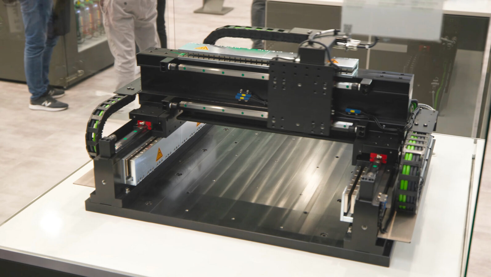 A large industrial 3D printer or CNC machine sits on a display table at a trade show, with visible mechanical parts and rails. People are standing in the background.