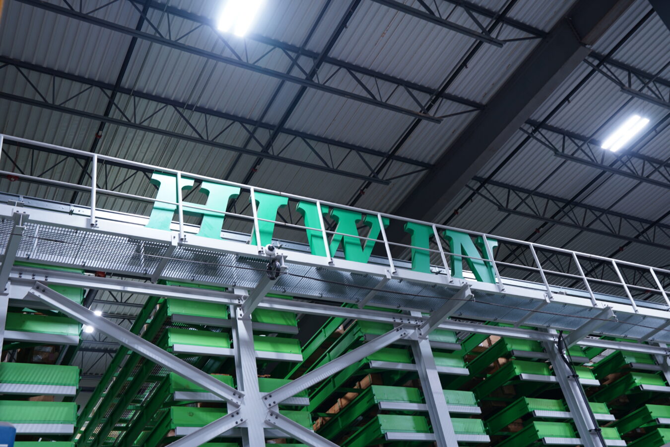 Large green letters spelling HIWIN are mounted above stacked green shelves inside an industrial warehouse with a high metal ceiling and overhead lights.