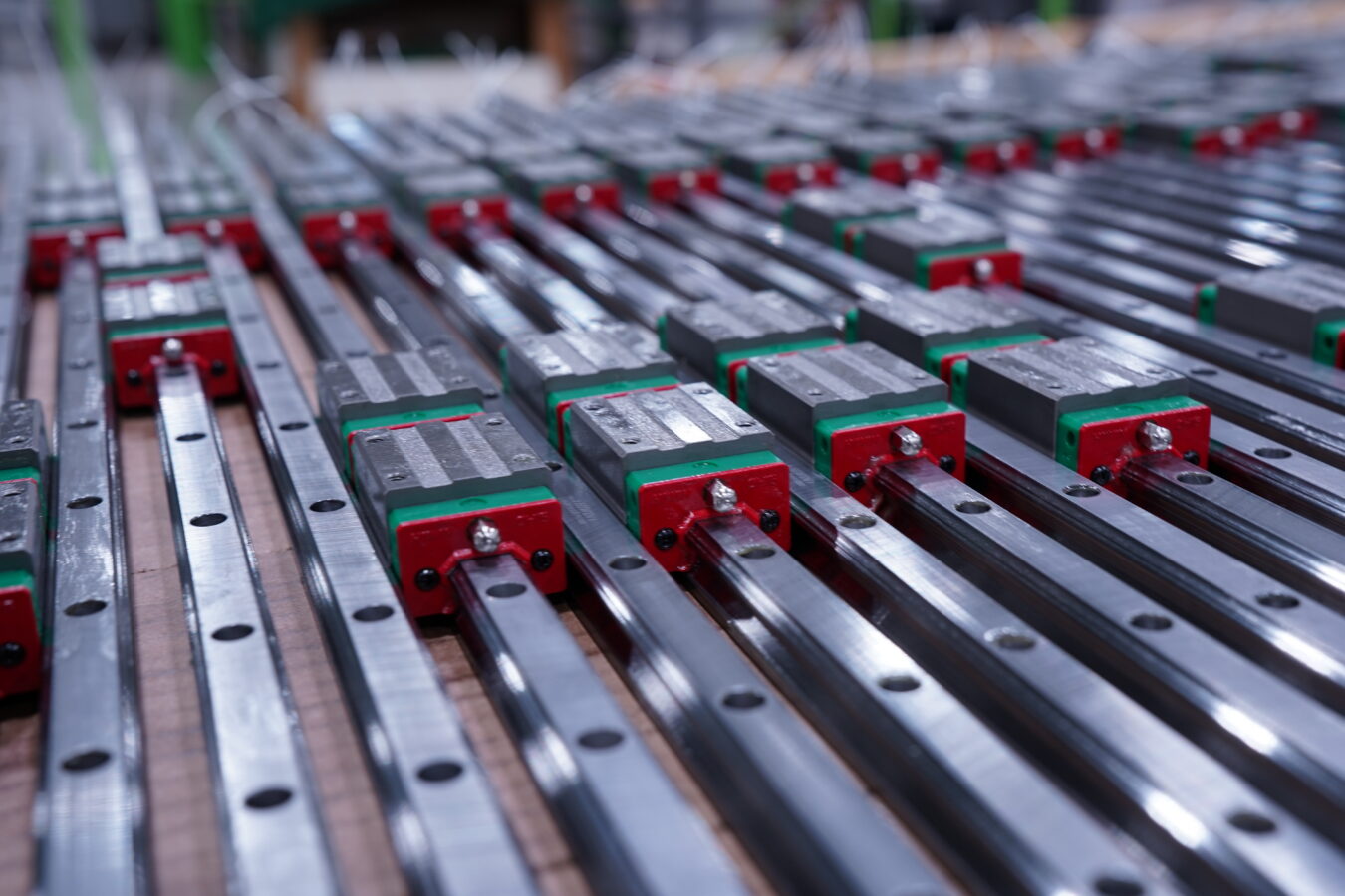 Several linear guide rails with red and green bearing blocks are aligned in parallel rows, reflecting light in an industrial manufacturing setting.
