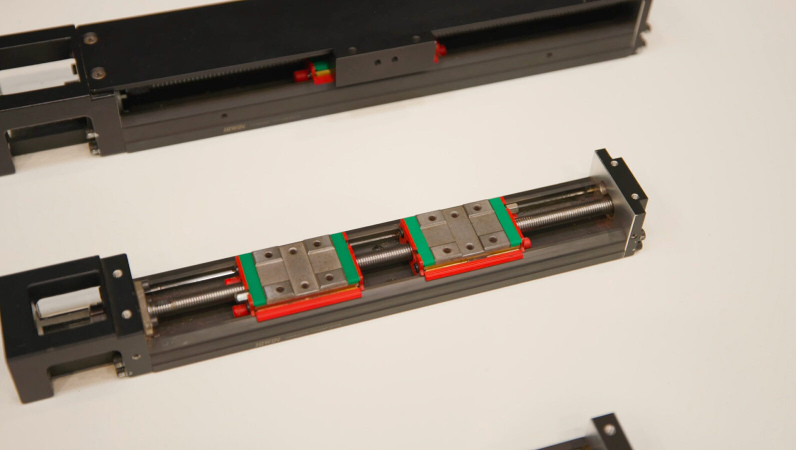 Metal linear rail with two green and red sliding carriages mounted on rods, used for precise motion in machinery, placed on a white surface.