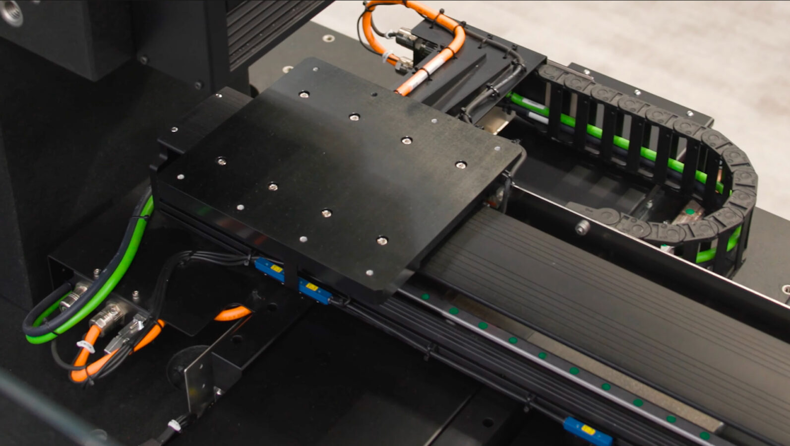Close-up of a black industrial machine with metal parts, cables, and green and orange wires, showing a moving platform and track system, likely for automation or manufacturing use.