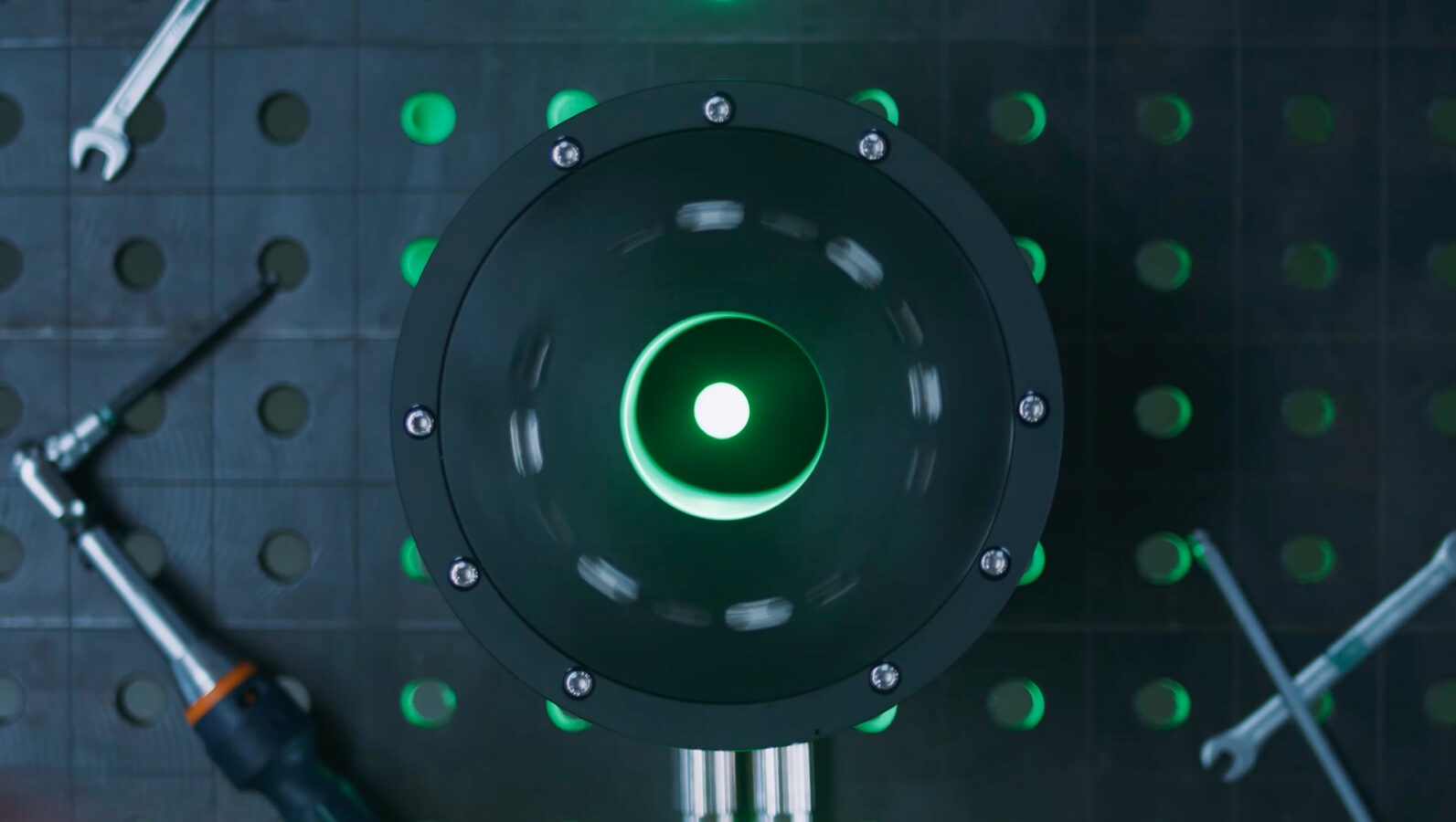 Top-down view of a circular black device with glowing green light inside, placed on a grid-patterned surface with green dots. Wrenches and a screwdriver are nearby.