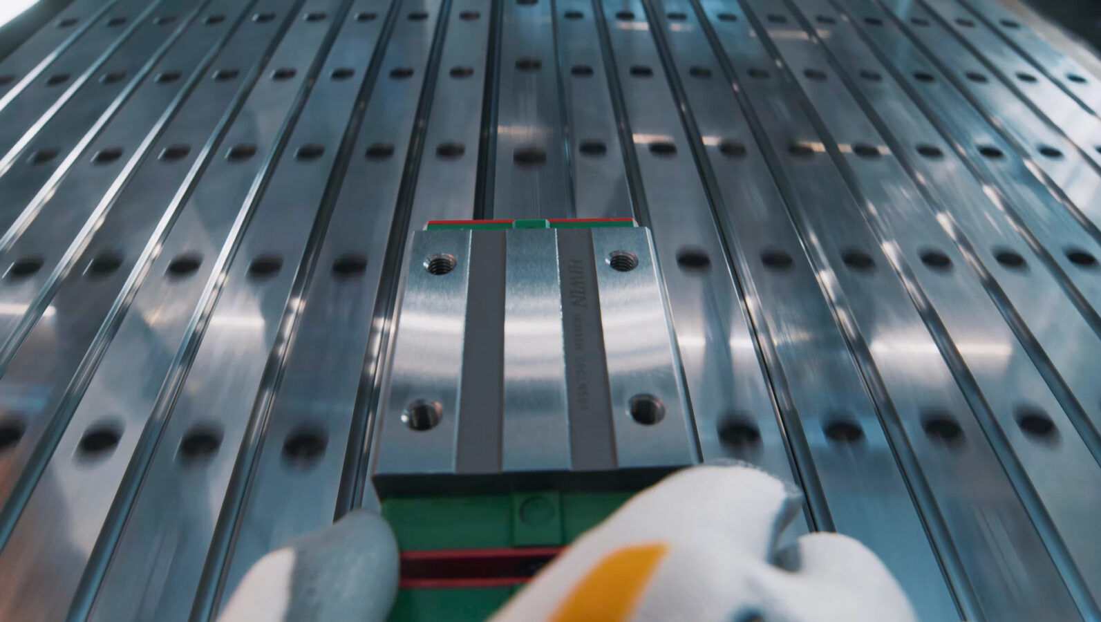 A gloved hand adjusts a metal linear rail block on a shiny, perforated metal surface, likely part of an industrial or manufacturing machine.