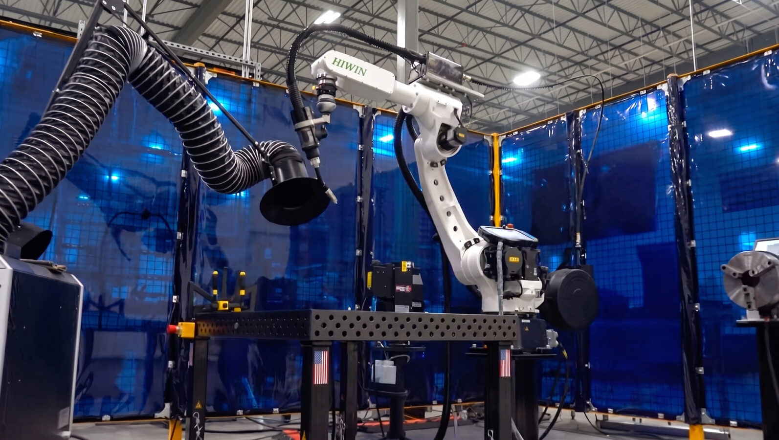 A white industrial robotic arm operates in a factory with blue safety barriers, metal fixtures, and ventilation hoses visible in the background.