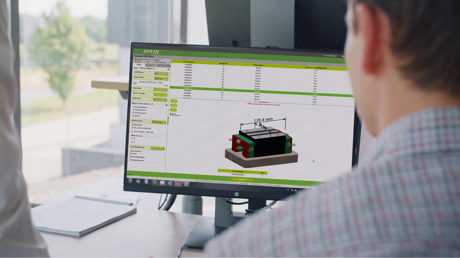 A person views a computer monitor displaying a 3D technical drawing with dimensions and specifications for a rectangular component. The interface features green and white colors, and there is a window in the background.