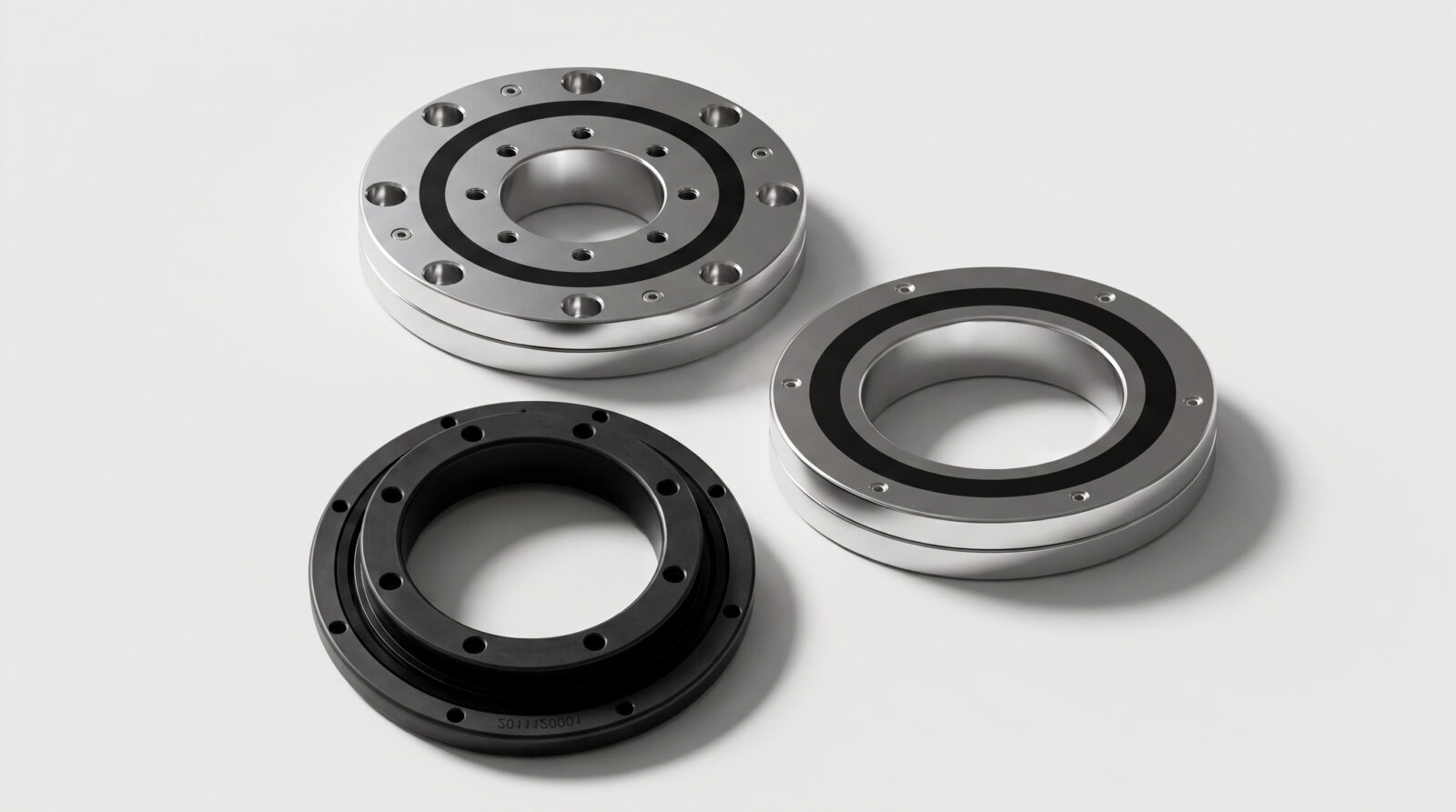 Three circular metal components, two silver and one black, with multiple holes and grooves, are arranged on a plain white surface. The objects appear to be mechanical parts, possibly bearings or housings.