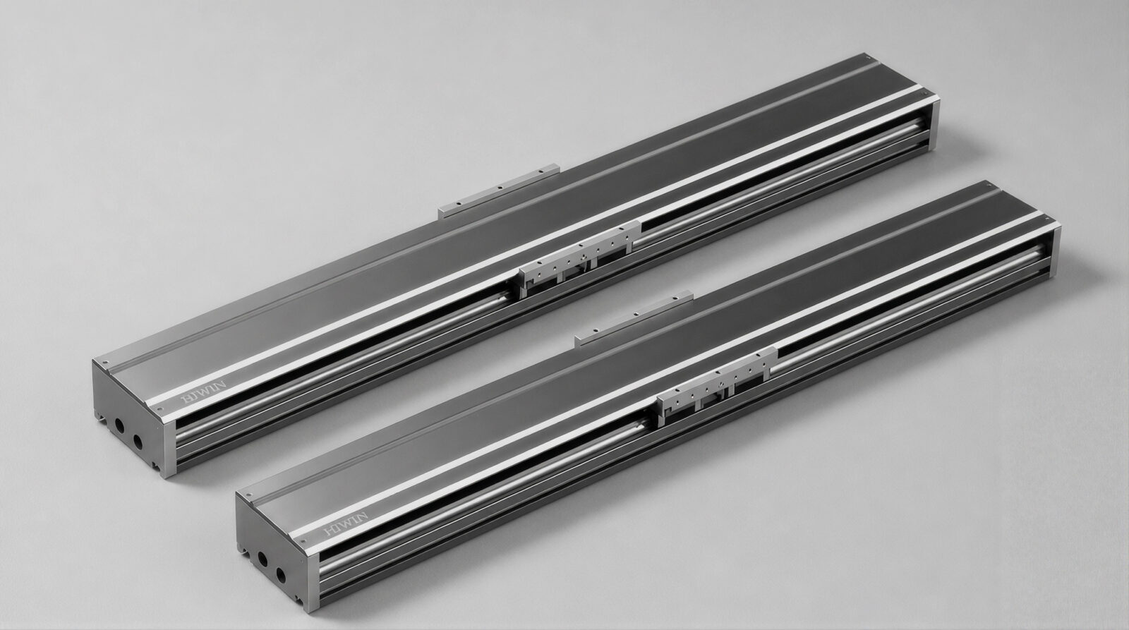 Two parallel metal linear guide rails with sliding carriages, placed on a plain gray background. The rails are silver with black details and appear to be industrial machine components.