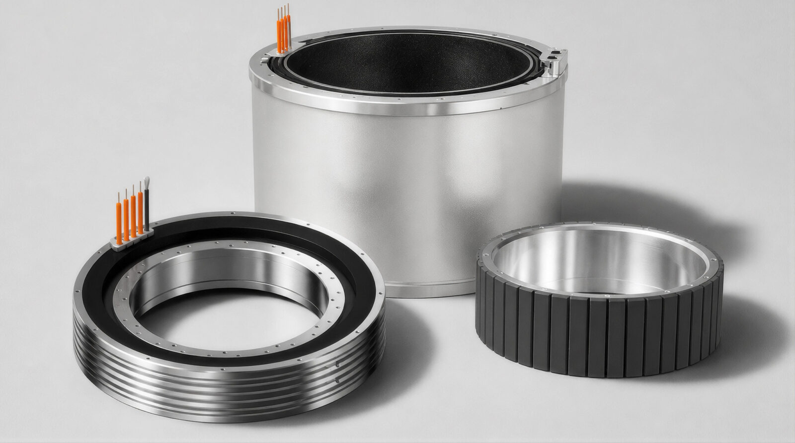 Three metallic cylindrical motor components, including a stator with orange wires, a rotor with black segments, and a large cylindrical housing, are arranged on a gray surface.