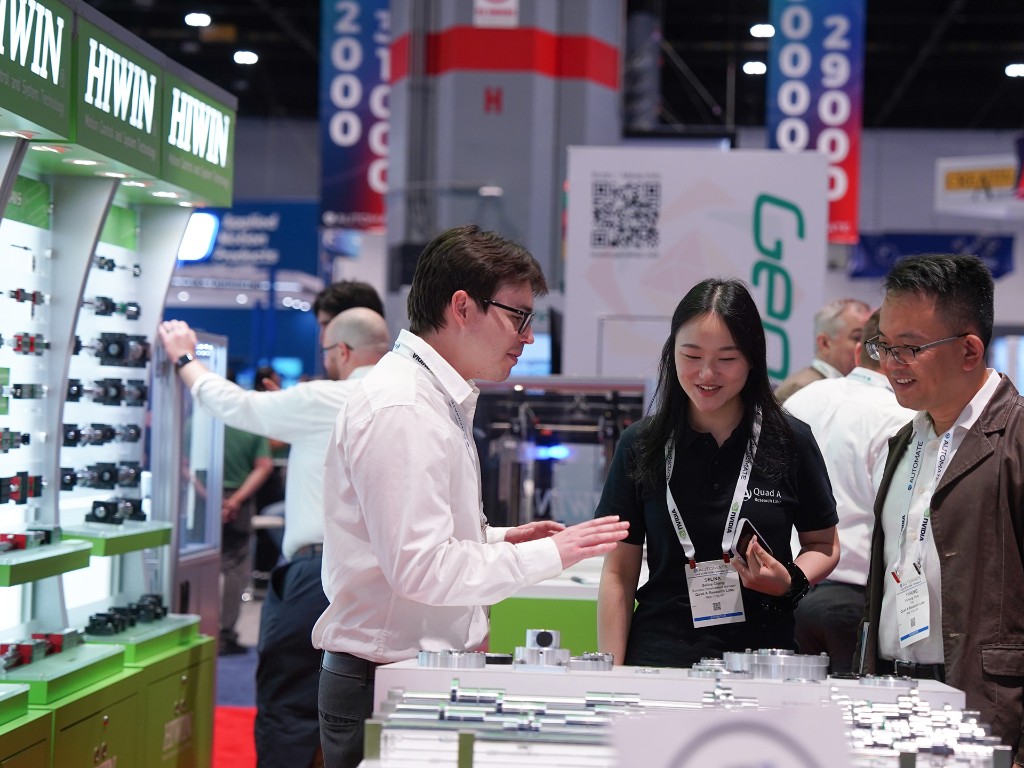 Three people talk at a trade show booth displaying mechanical parts. One man gestures while speaking to a woman and another man, both smiling. Exhibition banners and attendees are visible in the background.