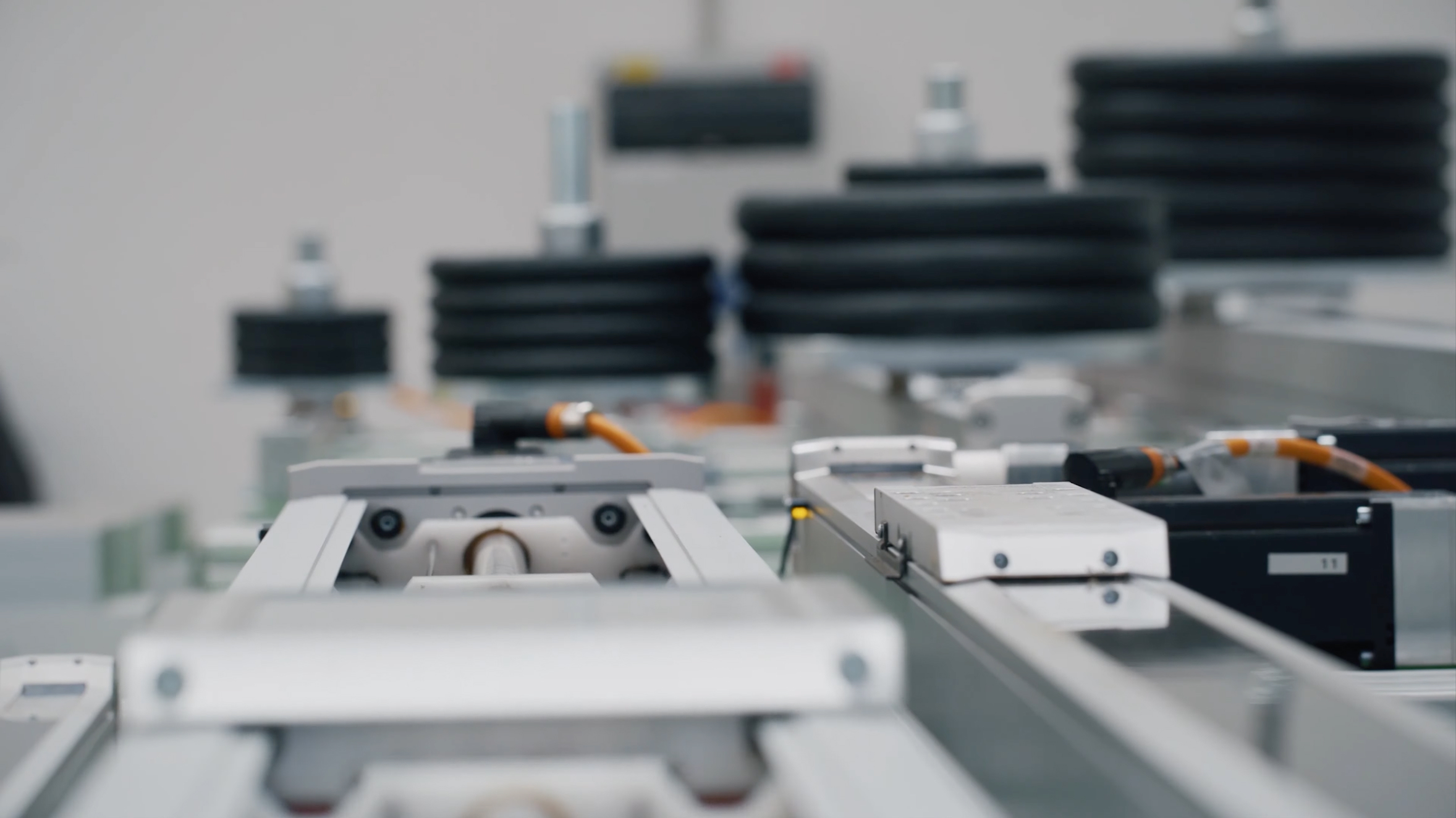 Close-up of industrial machinery components with metal rails, wires, and blurred black stacked objects in the background, suggesting a high-tech manufacturing or automation setting.