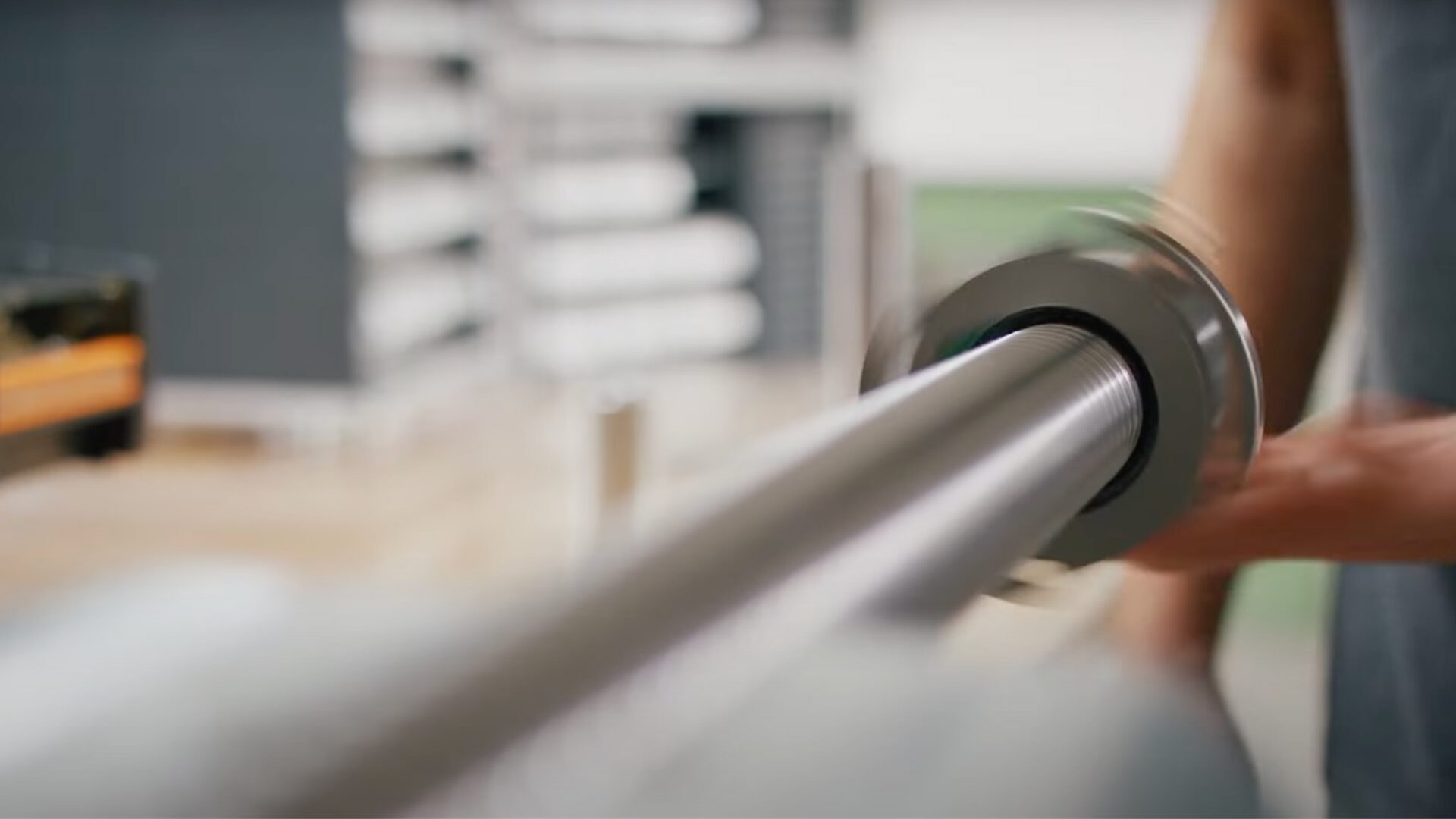 A persons hands install a metal bearing onto a long, cylindrical metal rod in a workshop or industrial setting. Shelves and equipment appear blurred in the background.