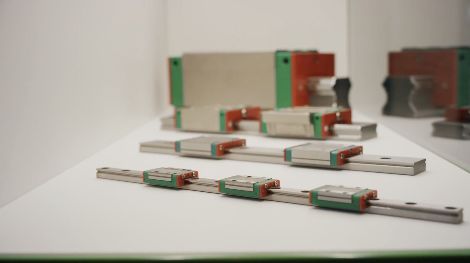 Several linear motion guide rails and blocks are displayed on a white surface, arranged in parallel rows, with metallic bodies and green and red end caps.