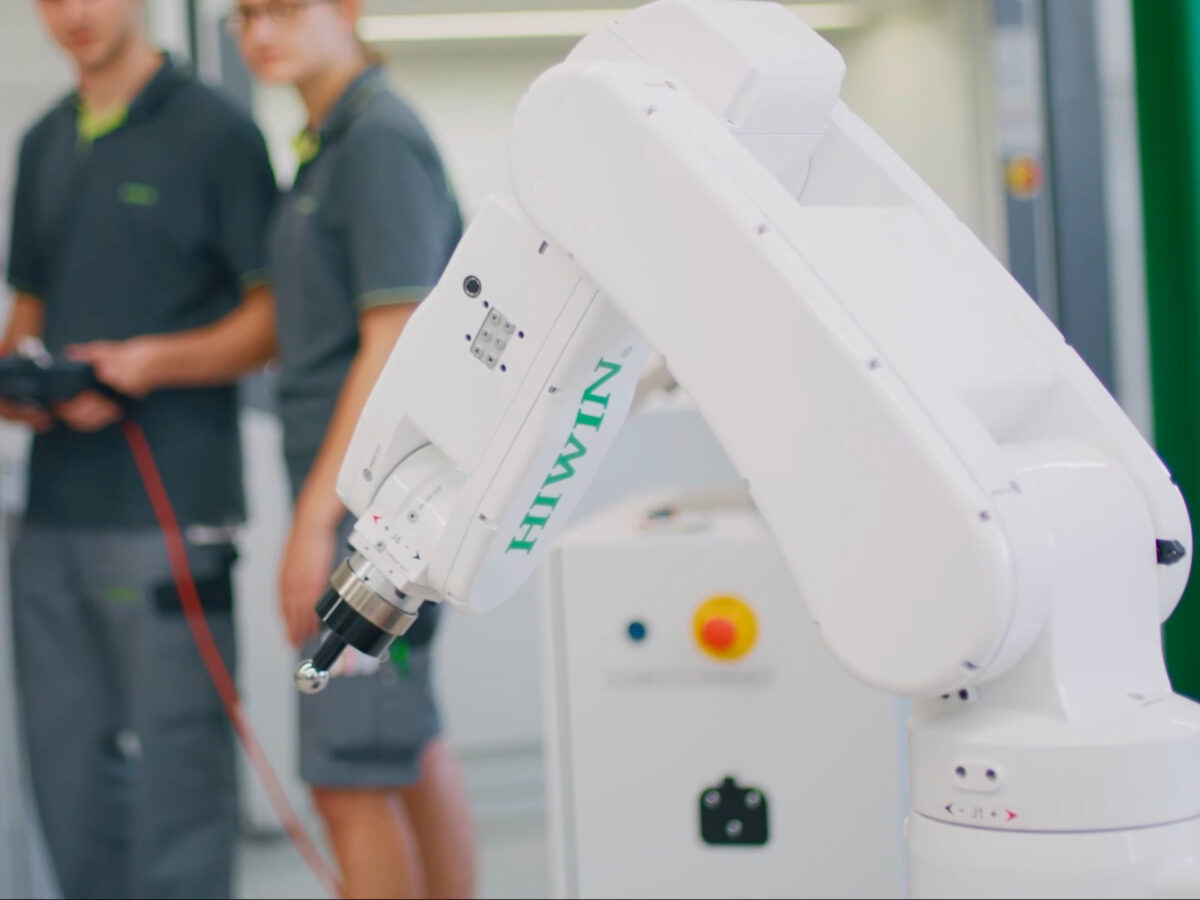 A white robotic arm in focus, with blurred figures of two people in grey uniforms and green trim in the background, appearing to operate or observe the machinery in a modern, bright setting.