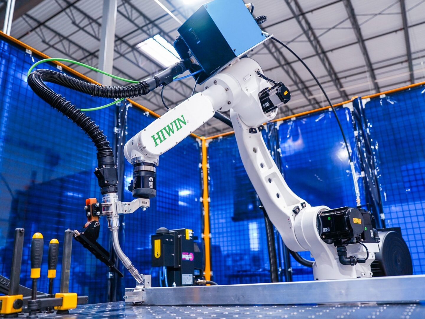 A white industrial robotic arm labeled HIWIN operates in a modern factory setting with blue safety barriers and various tools on a metal workbench.
