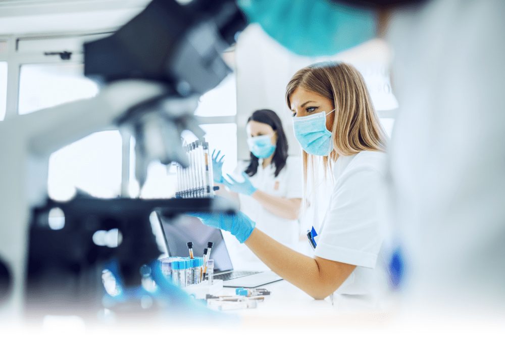 Two scientists in lab coats and masks work with microscopes and test tubes in a bright laboratory. One focuses on samples, while the other works at a laptop. Blue gloves and lab equipment are visible.