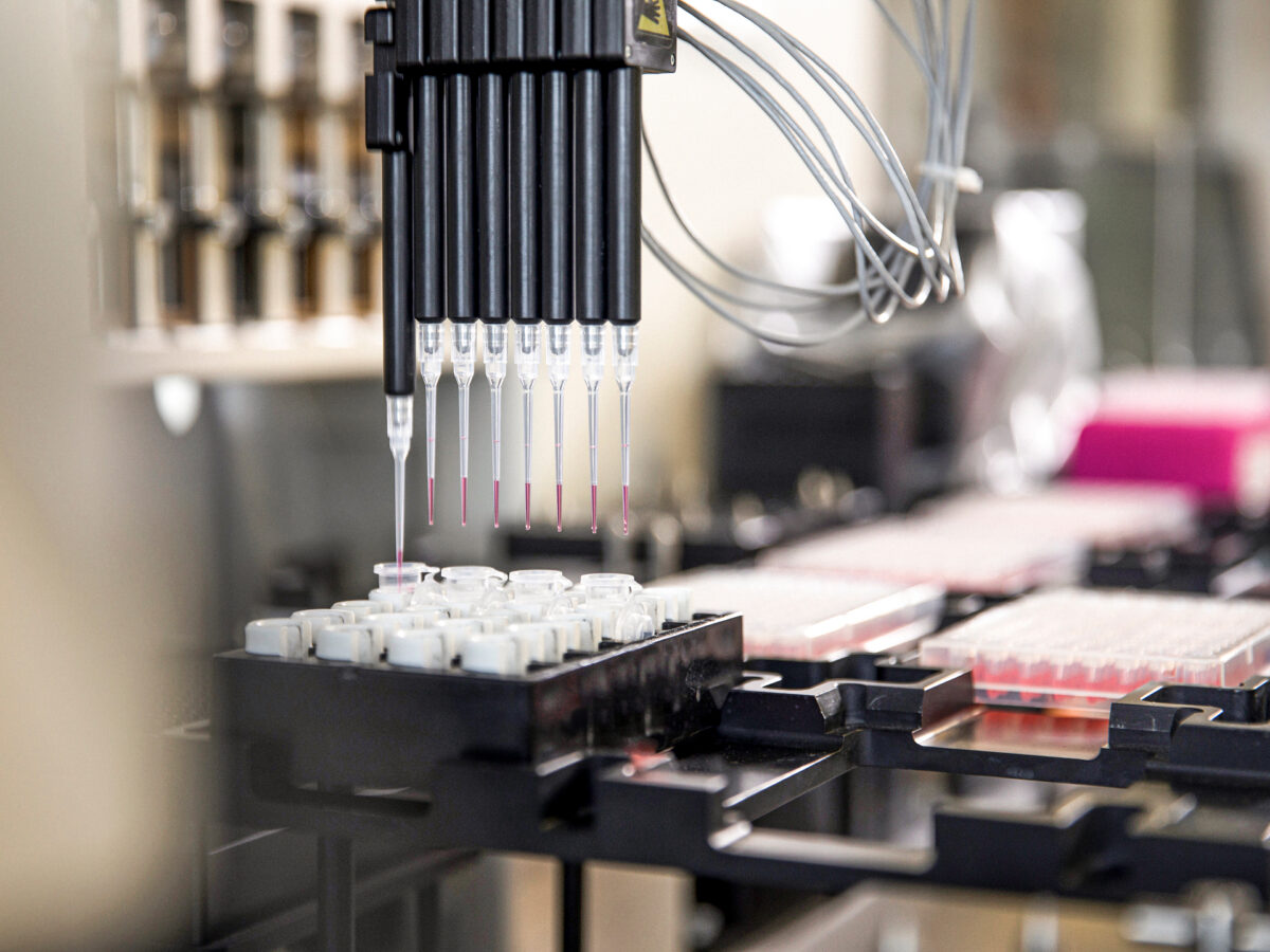 A close-up of an automated laboratory pipetting machine dispensing liquid into numerous small test tubes arranged in rows on a tray.