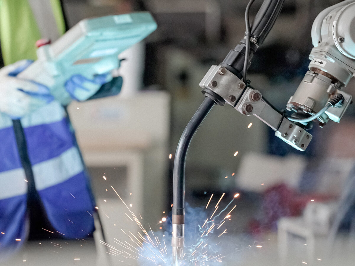 A robotic arm welds metal, producing sparks, while a person in safety gear operates a control device in the background.