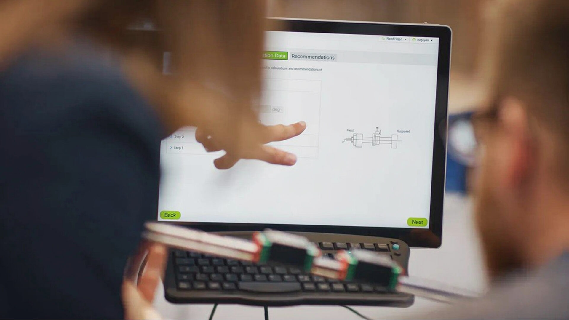Two people work at a computer showing a technical diagram. One person points at the screen, while the other holds an electronic device with visible sensors and wires in the foreground.
