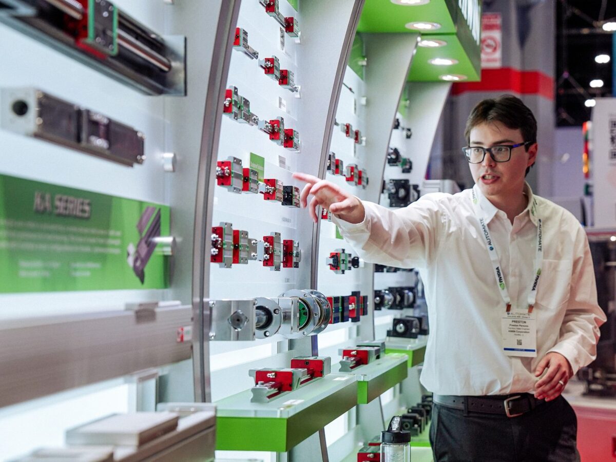 A person in glasses and a white shirt points at mechanical parts displayed on a wall at a trade show booth. The background features various small machines and informational signs.